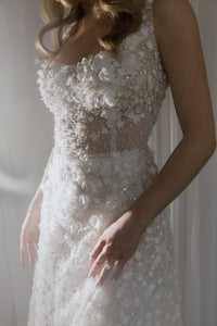 Dreamy semi-transparent wedding gown fully covered in sequins and 3D flower appliques. Featuring a square corset on straps, and an embellished maxi skirt with a delicate train.