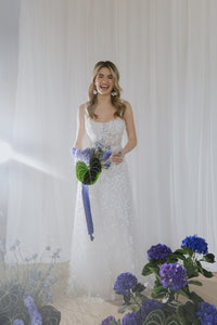 Dreamy semi-transparent wedding gown fully covered in sequins and 3D flower appliques. Featuring a square corset on straps, and an embellished maxi skirt with a delicate train.