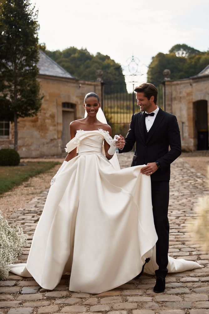 Chic off-the-shoulder mikado wedding gown. Featuring an artfully draped bodice adorned with 3D flower applique that emphasizes the neckline, an open back with a lace-up closure, and a long train.