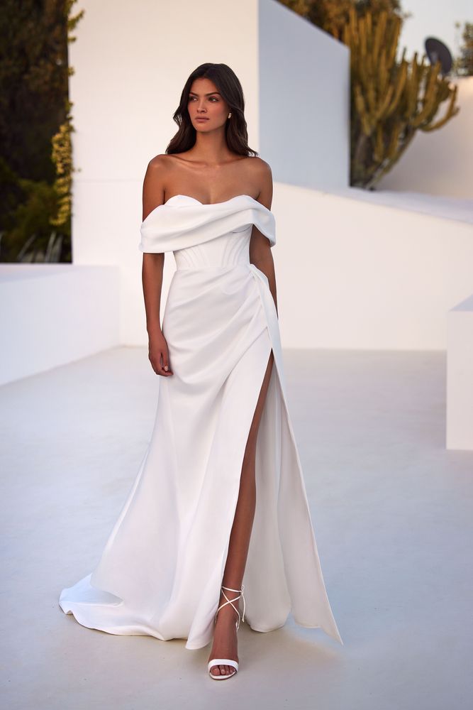 Chic off-the-shoulder wedding gown with a boned corset with originative integrated geometric cups that beautifully accentuate the neckline. The gown also boasts an open back with a lace-up closure or zipline by choice, a full skirt with a side slit, and a long train.