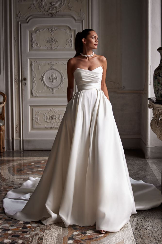 Elegant A-line wedding gown cut from mikado. The sleeveless design features a geometric neckline and a draped waist that flows into a gorgeous full skirt. Can be complemented with an off-the-shoulder mikado bolero with sheer long sleeves embellished with crystals.