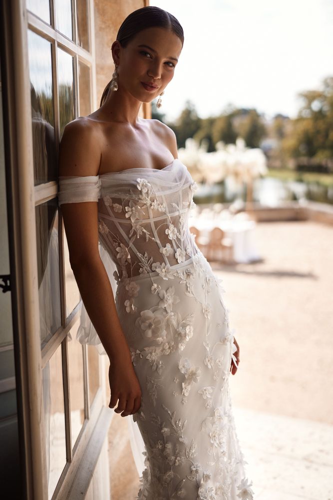 Romantic mermaid gown cut from lace with a semi-transparent sequined bodice with a straight neckline, 3D floral lace applique along the entire gown, off-the-shoulder tulle sleeves tied in the bow with wings on the back, and a short puffy tulle train.