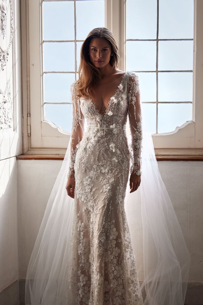 Delicate semi-transparent lace-adorned gown with 3D floral applique and silver shiny ornament. Featuring a plunging illusion V-neck, long sleeves, a buttoned back, and a nude lining skirt.
