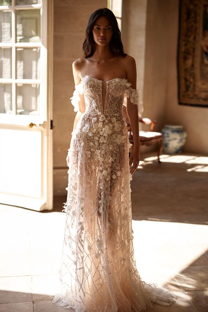 Mesmerizing semi-transparent shiny A-line wedding gown adorned with 3D flowers, sequins, beadings, and pearls. Featuring a sweetheart corset with a plunging neckline, off-the-shoulder tulle sleeves embellished with 3D flowers, lace-up closure on the back, and a short train.
