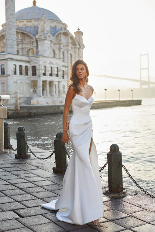 Feminine strapless satin wedding dress in white. Features a delicate heart-shaped neckline, draped skirt with a thing slit, and an open back with a lace-up closure. Complemented with detachable puffy sleeves and a long cathedral train.
